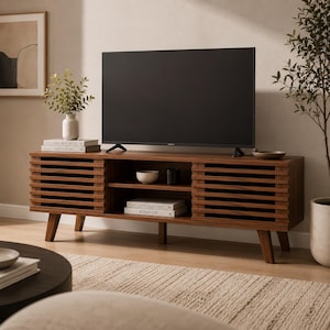 May include: A mid-century modern style TV stand in a warm brown finish. The stand features slatted sliding doors, open shelving, and angled legs. A large black television sits atop the stand, with decorative items and books on display.