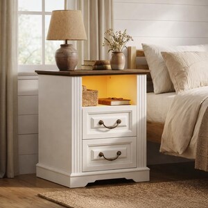May include: A white nightstand with two drawers and bronze-colored handles. The top is a dark wood, and a lamp with a tan shade sits on top. A small shelf is illuminated with a warm light, holding a woven basket and a book.