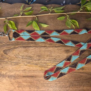 May include: A beaded lanyard with a silver-colored clasp. The lanyard features a geometric pattern of turquoise, red, black, and gold beads. The lanyard is lying on a wooden surface.