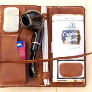 May include: A brown leather pipe tobacco pouch with a pipe, a lighter, a pack of tobacco, and a cleaning tool. The tobacco pack is labeled "Captain Black" and the pouch has a strap.