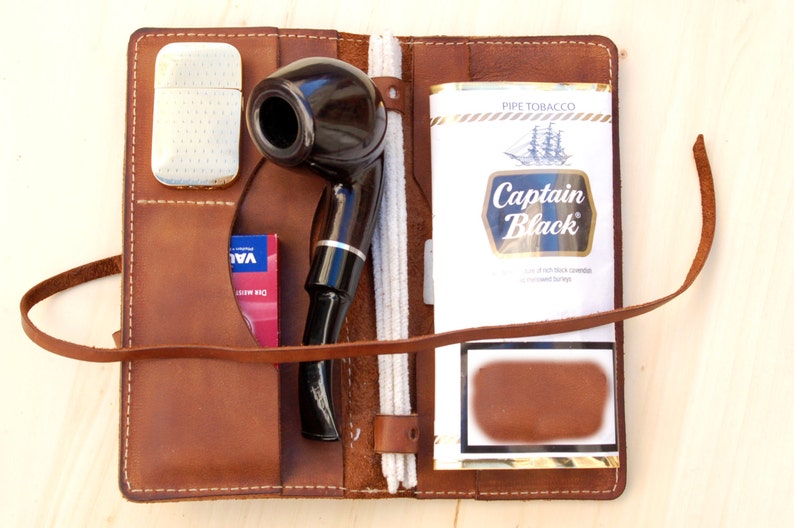 May include: A brown leather pipe tobacco pouch with a pipe, a lighter, a pack of tobacco, and a cleaning tool. The tobacco pack is labeled "Captain Black" and the pouch has a strap.
