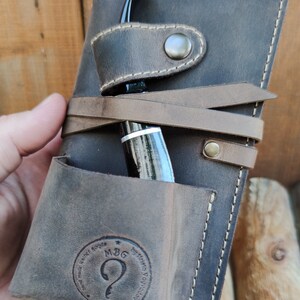 May include: A brown leather pipe case with a pocket for a pipe and a strap to secure it. The case has a stitched border and a logo that reads "MBG by Hanna Novicova est. 2005".