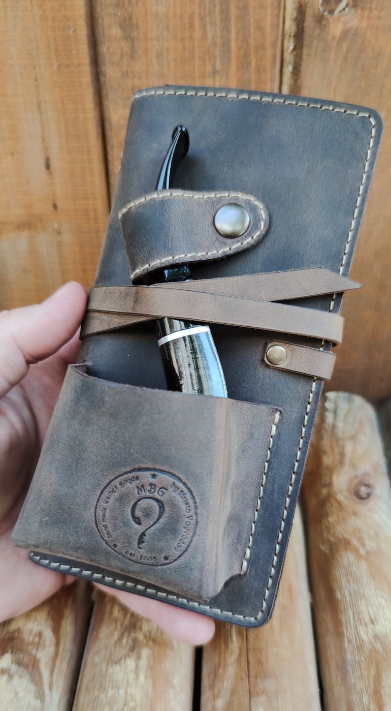 May include: A brown leather pipe case with a pocket for a pipe and a strap to secure it. The case has a stitched border and a logo that reads "MBG by Hanna Novicova est. 2005".