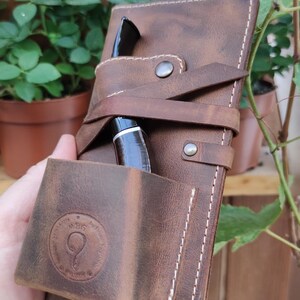 May include: A brown leather pipe case with a strap and a pocket for a pipe. The case has a white stitching detail and a circular logo with the text "MBG" and a stylized letter "Q".