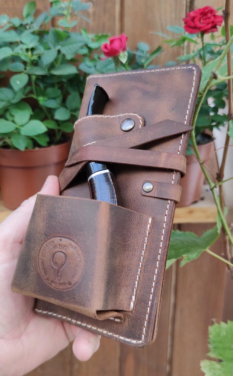 May include: A brown leather pipe case with a strap and a pocket for a pipe. The case has a white stitching detail and a circular logo with the text "MBG" and a stylized letter "Q".