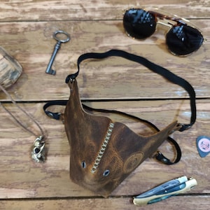 Pode incluir: Uma m&aacute;scara facial de couro marrom com uma al&ccedil;a, detalhes de costura e dois orif&iacute;cios para os olhos. A m&aacute;scara est&aacute; sobre uma superf&iacute;cie de madeira com uma chave, &oacute;culos de sol, um colar e um palheta de guitarra.