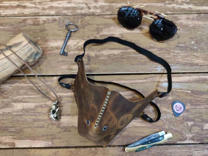 Pode incluir: Uma m&aacute;scara facial de couro marrom com uma al&ccedil;a, detalhes de costura e dois orif&iacute;cios para os olhos. A m&aacute;scara est&aacute; sobre uma superf&iacute;cie de madeira com uma chave, &oacute;culos de sol, um colar e um palheta de guitarra.