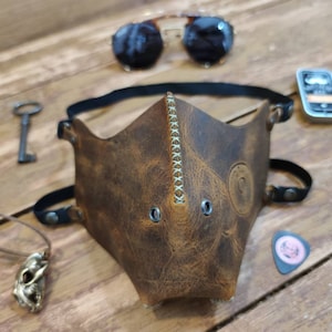 Pode incluir: Uma m&aacute;scara facial de couro marrom com um design costurado e ilh&oacute;s de metal. A m&aacute;scara tem tiras ajust&aacute;veis e &eacute; feita de couro envelhecido.