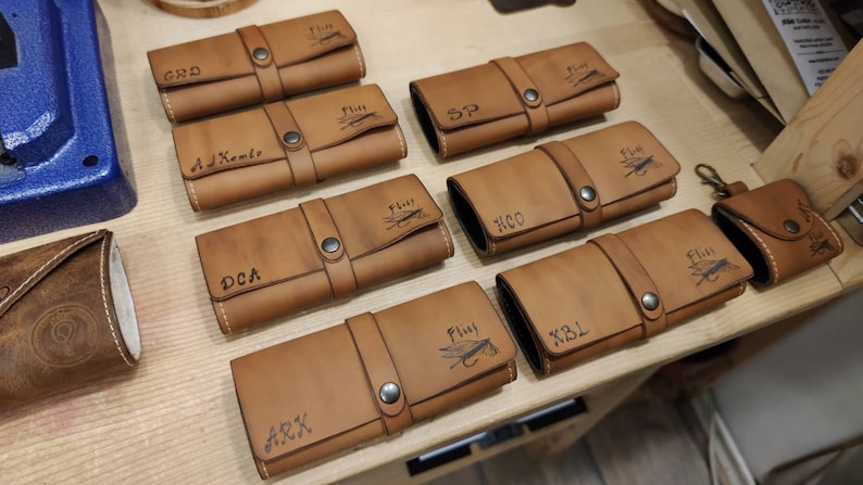 May include: A collection of ten brown leather fly fishing cases with various names and initials engraved on them. Each case has a flap with a snap closure and a fly fishing lure design.