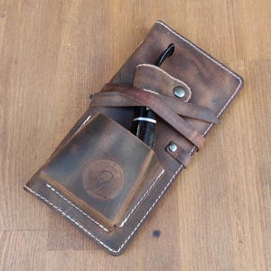 May include: A brown leather pipe case with a stitched design and a small pocket for a pipe. The case has a brown leather strap with a metal buckle.