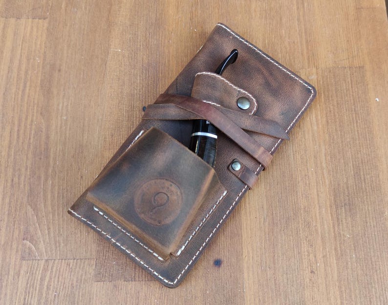 May include: A brown leather pipe case with a stitched design and a small pocket for a pipe. The case has a brown leather strap with a metal buckle.