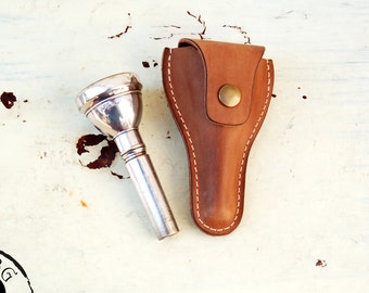 May include: A silver trumpet mouthpiece with a leather case. The case has a brown leather strap and a brass button.