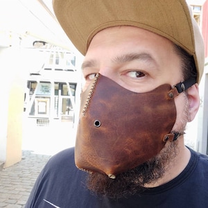 Pode incluir: Uma m&aacute;scara facial de couro marrom com uma borda costurada e um pequeno furo no centro. A m&aacute;scara &eacute; usada por uma pessoa com barba e um bon&eacute; de beisebol marrom.