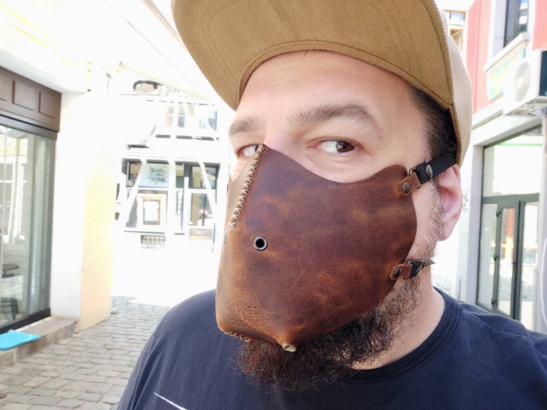 Pode incluir: Uma m&aacute;scara facial de couro marrom com uma borda costurada e um pequeno furo no centro. A m&aacute;scara &eacute; usada por uma pessoa com barba e um bon&eacute; de beisebol marrom.