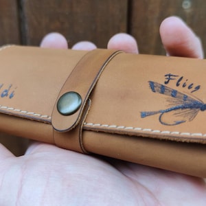 May include: A brown leather fly fishing fly box with the name "Maddi" and the word "Fly" engraved on the front. The box has a white fleece lining and a brown leather strap with a metal button.