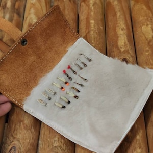 May include: A brown leather fly fishing fly box with a white fur interior. The box is open and shows a variety of flies.