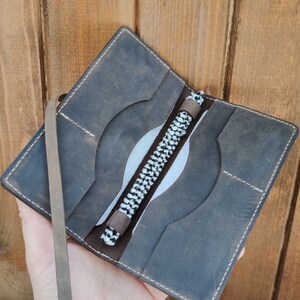 May include: A brown leather wallet with a white and black braided strap. The wallet has multiple pockets and a flap closure.