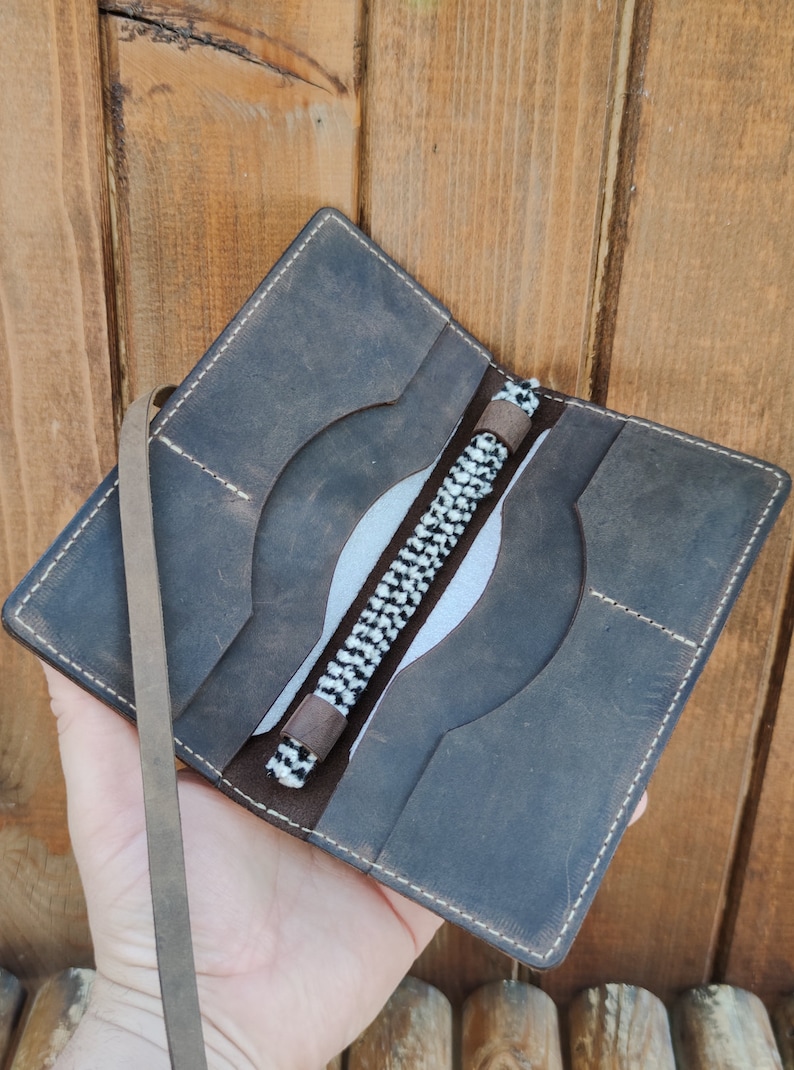 May include: A brown leather wallet with a white and black braided strap. The wallet has multiple pockets and a flap closure.
