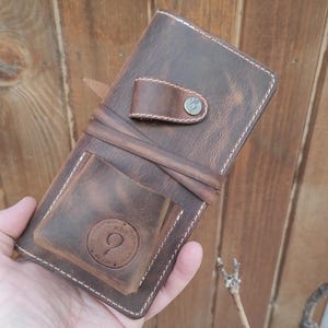 May include: A brown leather wallet with a stitched edge and a metal button. The wallet has a flap with a small pocket and a larger pocket with a circular design.