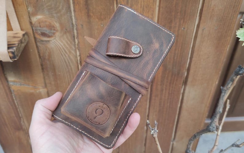 May include: A brown leather wallet with a stitched edge and a metal button. The wallet has a flap with a small pocket and a larger pocket with a circular design.