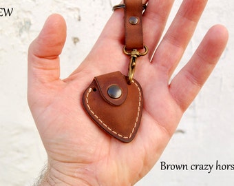 May include: A brown leather guitar pick holder with a snap closure and a key ring. The holder is stitched with white thread and has a distressed leather finish. "NEW" and "Brown crazy horse" are written in the image.