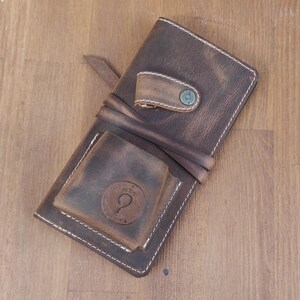 May include: A brown leather wallet with a flap closure and a small pocket on the front. The wallet has a distressed look and white stitching. The wallet has a metal snap closure and a metal loop on the front.