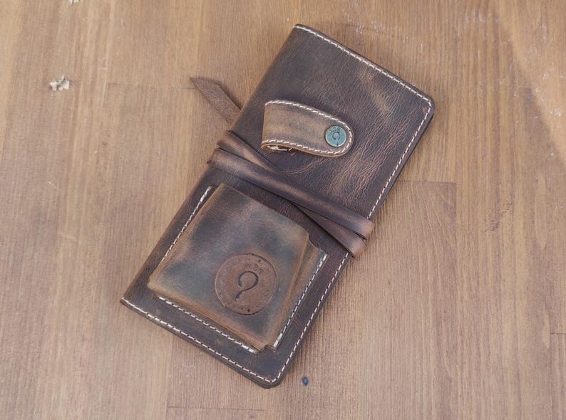 May include: A brown leather wallet with a flap closure and a small pocket on the front. The wallet has a distressed look and white stitching. The wallet has a metal snap closure and a metal loop on the front.