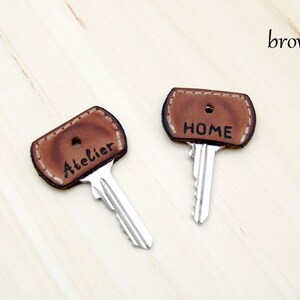 May include: Two brown leather key covers with stitched edges. The covers are labeled "HOME" and "Atelier".