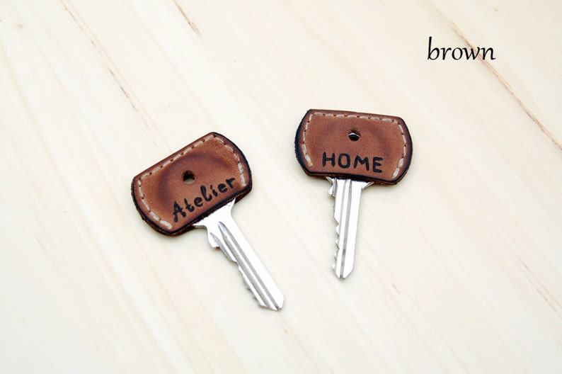 May include: Two brown leather key covers with stitched edges. The covers are labeled "HOME" and "Atelier".