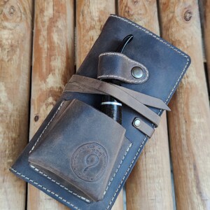 May include: A brown leather pipe case with a strap and a pocket for a pipe. The case has a stitched design and a logo that says "Handmade Goods by Quinn Design".