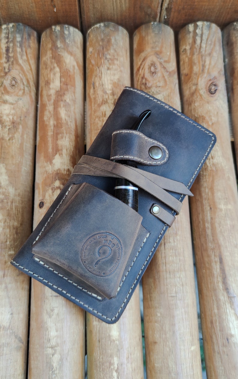 May include: A brown leather pipe case with a strap and a pocket for a pipe. The case has a stitched design and a logo that says "Handmade Goods by Quinn Design".