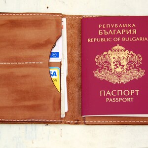 May include: A brown leather passport holder with a burgundy Bulgarian passport inside. The passport has the text "Република България" and "REPUBLIC OF BULGARIA" on the cover, along with the Bulgarian coat of arms. The passport holder has a pocket for cards and a flap to secure the passport.