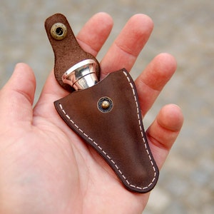 May include: A brown leather pouch with a white stitch detail and a silver metal flask inside. The pouch has a flap with a snap closure and a decorative metal button.