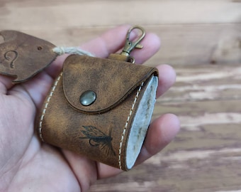 May include: A brown leather fly fishing lure box with a fish-shaped leather keychain attached. The box has a white stitching detail and a small metal button closure. The keychain has a small metal loop for attaching to a belt or backpack.