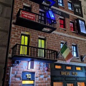 May include: A detailed miniature of a New York City street scene, featuring brick buildings with balconies, illuminated windows, and a "Brew Pub" sign. Includes an "Apartment For Sale" sign and an Irish flag.