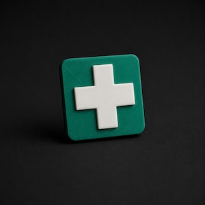 Op de afbeelding: Een vierkant embleem met een effen wit kruis gecentreerd op een donkergroene achtergrond. Het object heeft een matte, getextureerde afwerking met licht afgeronde hoeken, wat zorgt voor een strak, geometrisch ontwerp met een verhoogd, driedimensionaal uiterlijk.