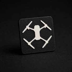 May include: A square black plaque featuring a white, stylized graphic of a quadcopter drone centered on its surface. The drone icon displays a central body with four extended arms and propellers, creating a high-contrast design on the matte black base.