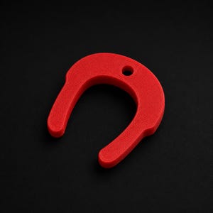 May include: A bright red, U-shaped plastic component featuring a textured surface finish and a single circular hole positioned at the top center of the arch. The object has two parallel downward-extending arms and a curved top section.