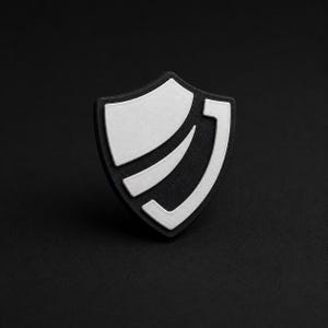 Puede incluir: Un emblema en forma de escudo con una base negra mate y detalles blancos en relieve. El diseño incluye una sección superior blanca sólida y dos franjas blancas curvas que siguen el contorno del escudo, creando un aspecto de alto contraste.