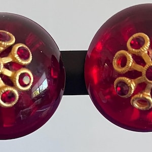 May include: A pair of round, deep red translucent cabochon earrings. Each earring features a central gold-toned metal floral-shaped ornament adorned with small, circular red rhinestones set into each petal and the center of the flower design.