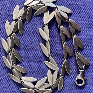 May include: A silver-toned metal necklace featuring a repeating pattern of small, smooth, leaf-shaped links connected in a continuous chain. The necklace includes a circular lobster claw clasp at one end, designed to secure the piece around the neck.
