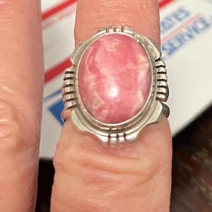 May include: A silver ring featuring an oval, pink gemstone with white marbling. The ring has a decorative, ridged design on the sides and a detailed setting around the stone. The ring is a piece of jewelry.