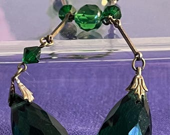 Puede incluir: Un par de pendientes colgantes con cuentas de cristal verde esmeralda facetadas y grandes colgantes en forma de lágrima. Los cristales están conectados por finas barras metálicas de tono antiguo y anillas, con tapas decorativas sobre cada lágrima.