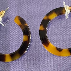 May include: A pair of hoop earrings featuring a classic tortoiseshell pattern in shades of dark brown and amber. The earrings are circular in shape with a smooth, polished finish and include gold-tone metal posts with clear plastic earring backs.