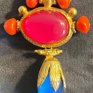 May include: A vintage brooch with a gold-tone metal setting. It features a large, oval, magenta-colored stone at the center, surrounded by orange accents. A blue teardrop-shaped stone hangs below, with gold leaf detailing. Additional orange accents are present.
