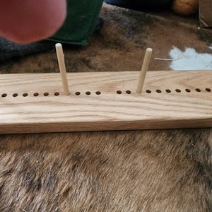 May include: A light-colored wooden yarn swift with four wooden pegs inserted. The swift has a rectangular base with evenly spaced holes for the pegs. The wood grain is visible, and the swift is on a textured surface.