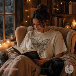 A woman sits in an armchair by a window, reading a book and wearing a cream-colored t-shirt with a dark celestial moth design.