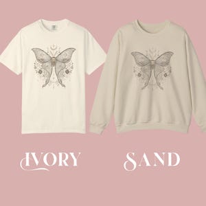 An ivory short-sleeved t-shirt and a sand long-sleeved sweatshirt, both displaying a gray and white moth design with crescent moons and floral accents.