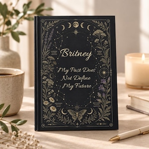 May include: A black hardcover journal featuring gold foil botanical illustrations, moon phases, and constellations. The cover displays the name Britney and the text My Past Does Not Define My Future, surrounded by lavender, yarrow, chamomile, and bellflower.