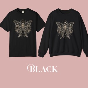 A black short-sleeved t-shirt and a black crewneck sweatshirt, both featuring a gold celestial moth design, are displayed side-by-side.